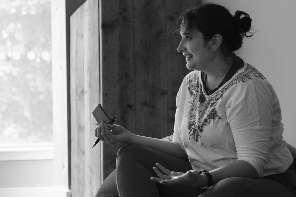 Laura is seated on a floor cushion. In the background, sunlight spills in through a large window. She is gesturing with sticky notes and a pen in hand. She is smiling with a bright, excited expression. 