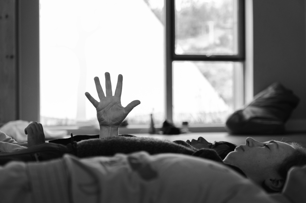 Three performers lie close together, their bodies are horizontal across the image. Their hands are rising, uncurling, and one hand shines upwards, fingers stretching out. In the background, sunlight spills through a large window.