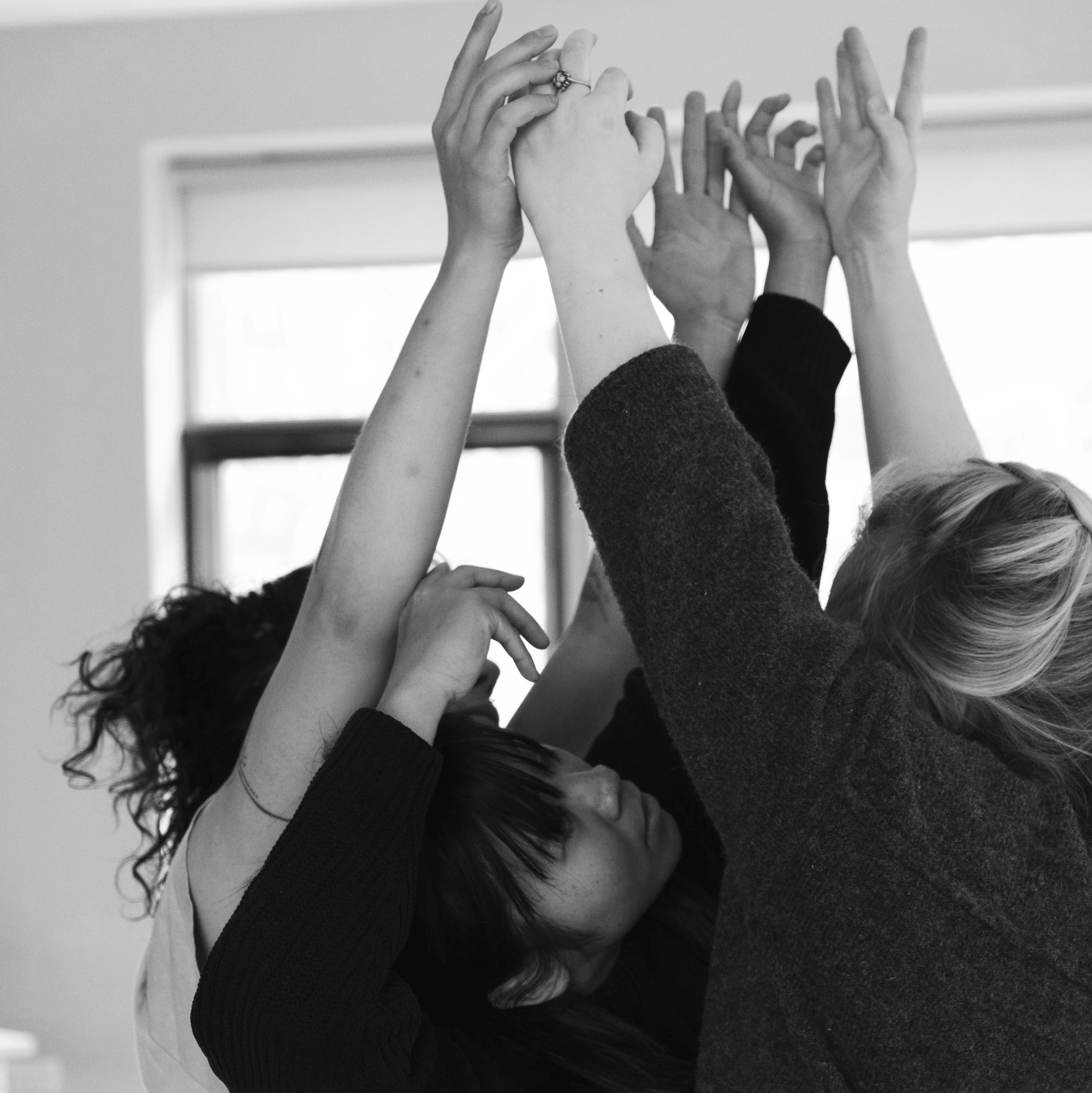 Three women performers are crowded together, their bodies close, reaching their arms upwards, their gaze follows. In the background, sunlight shines through large windows.