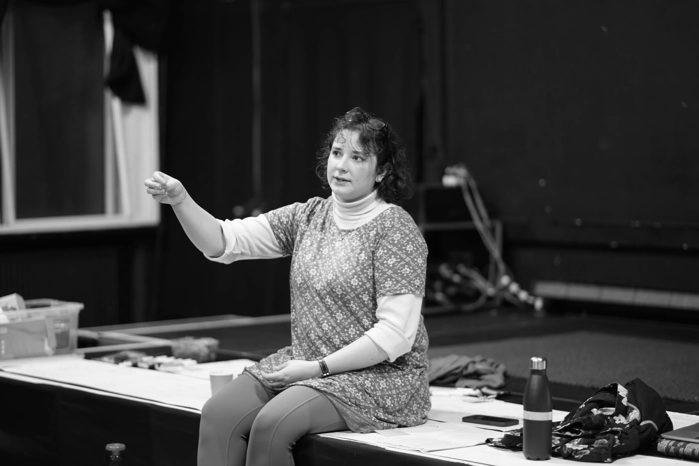 Laura sits on the edge of a stage surrounded by a large sheet of paper which has been annotated. She is gesturing with a thoughtful expression. 