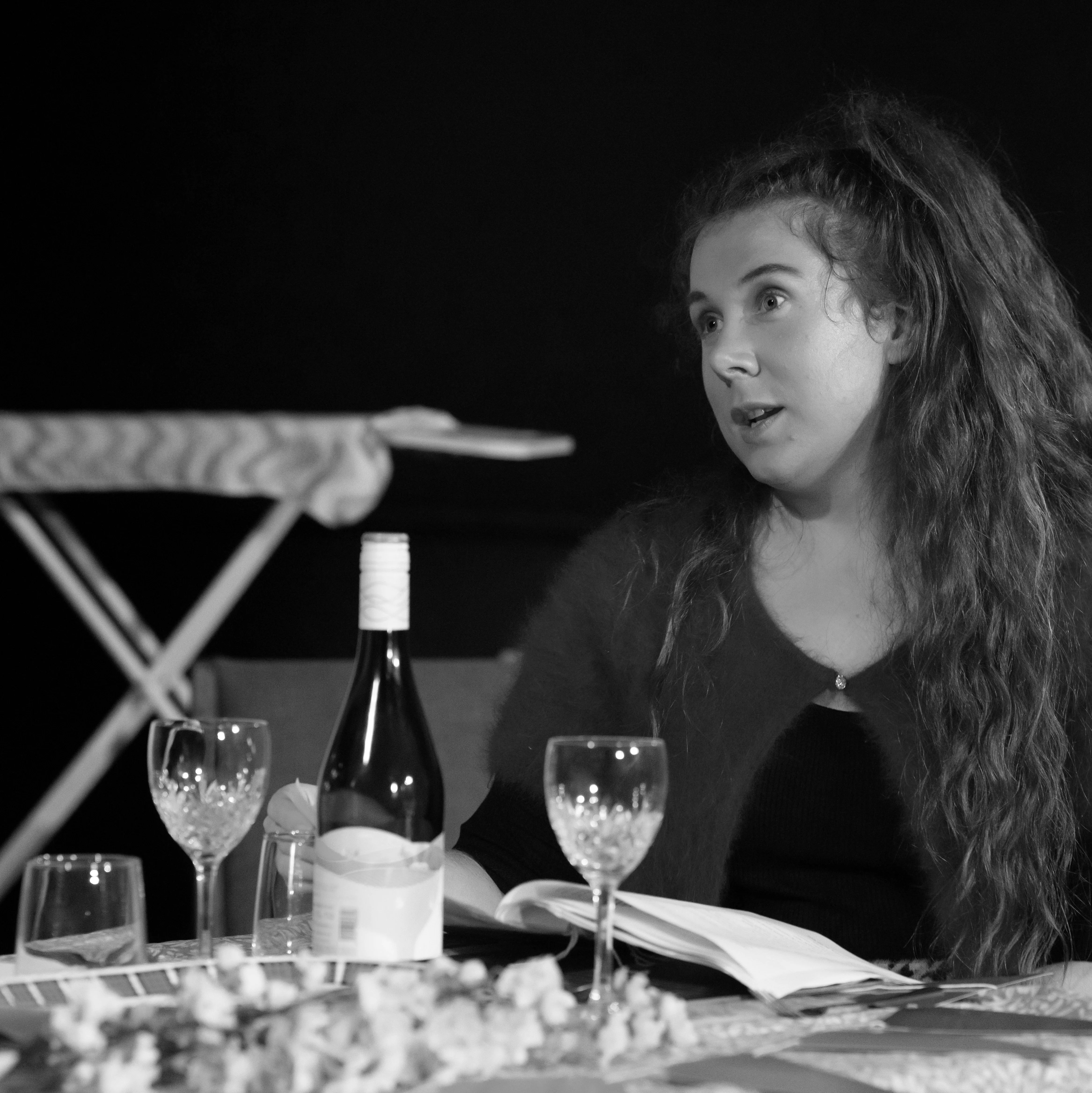 A woman actor sits at a dining table set with for a meal with a bottle of wine. Her script is in front of her and she looks out of the frame with an exasperated expression.
