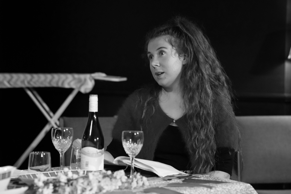 A woman actor sits at a dining table set with for a meal with a bottle of wine. Her script is in front of her and she looks out of the frame with an exasperated expression.