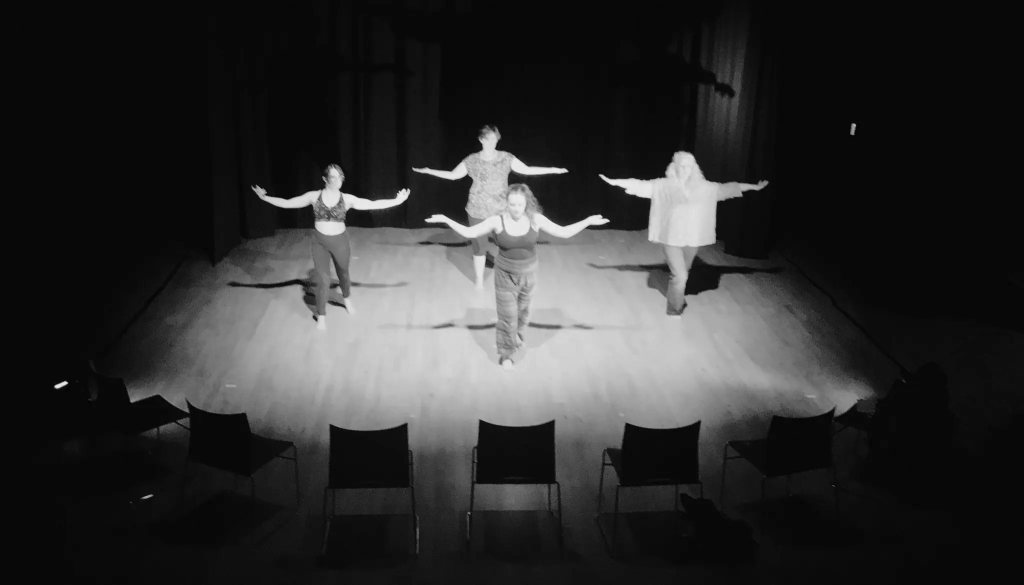 Four women stand on the stage. Seats are silhouetted in the foreground. The women are all performing the same movement, with arms outstretched and reaching upwards.