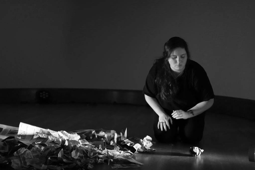 A woman performer kneels on the right, she is tentatively reaching towards a ball of paper. To the left is a pile of scrunched and torn newspaper pages. Lights cast deep shadows across them.