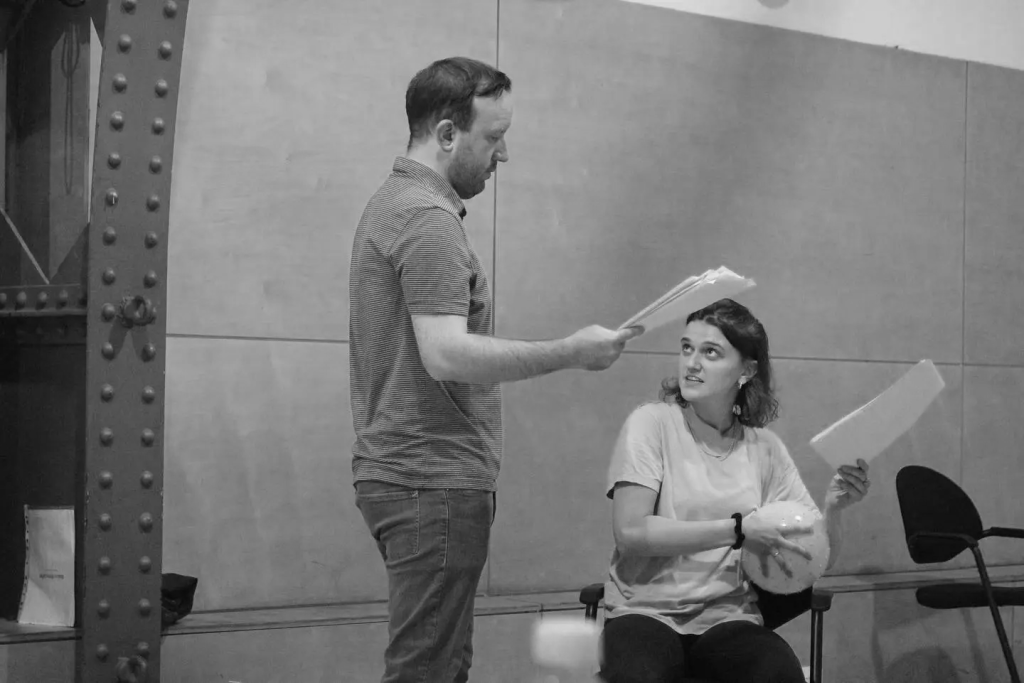 Two actors reading from their scripts; the man stands over the woman seated. The woman is talking to him with a playful expression. She is clutching a sports ball. 
