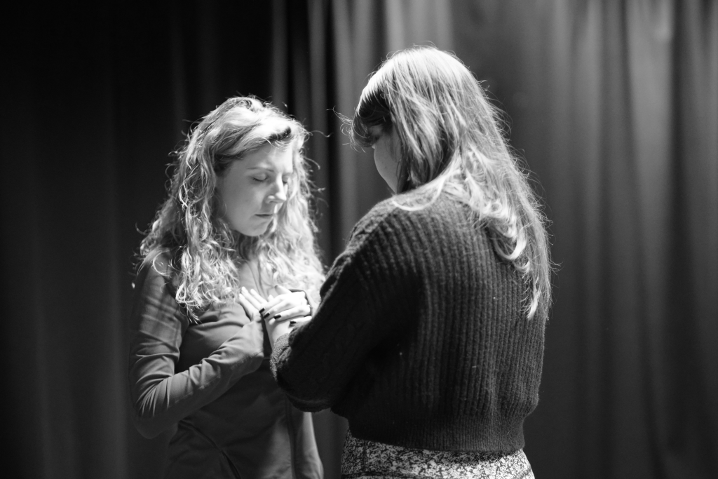 Two women performers face each other. They both hold their hands to one's heart-space. Their heads and bowed and eyes closed. Their energy is compassionate and peaceful. A stage curtain ripples in the background.